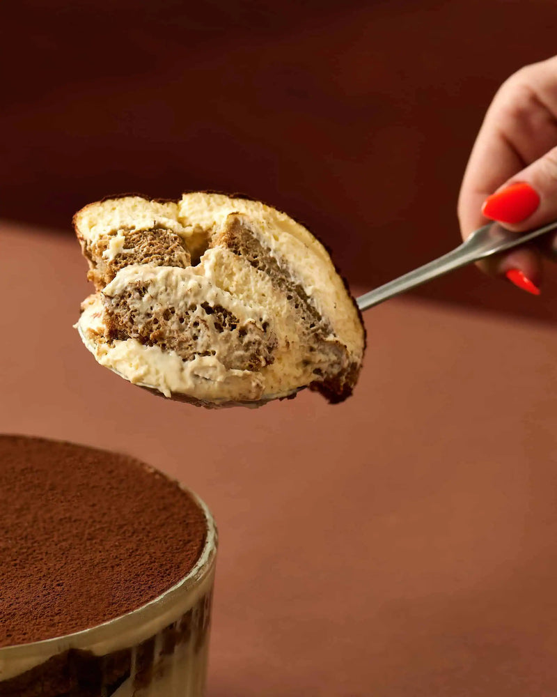 A close-up of a hand with bright red nail polish holding a slice of layered tiramisu on a metal spoon, with a partially visible cup of cocoa-dusted dessert in the foreground against a warm brown background.