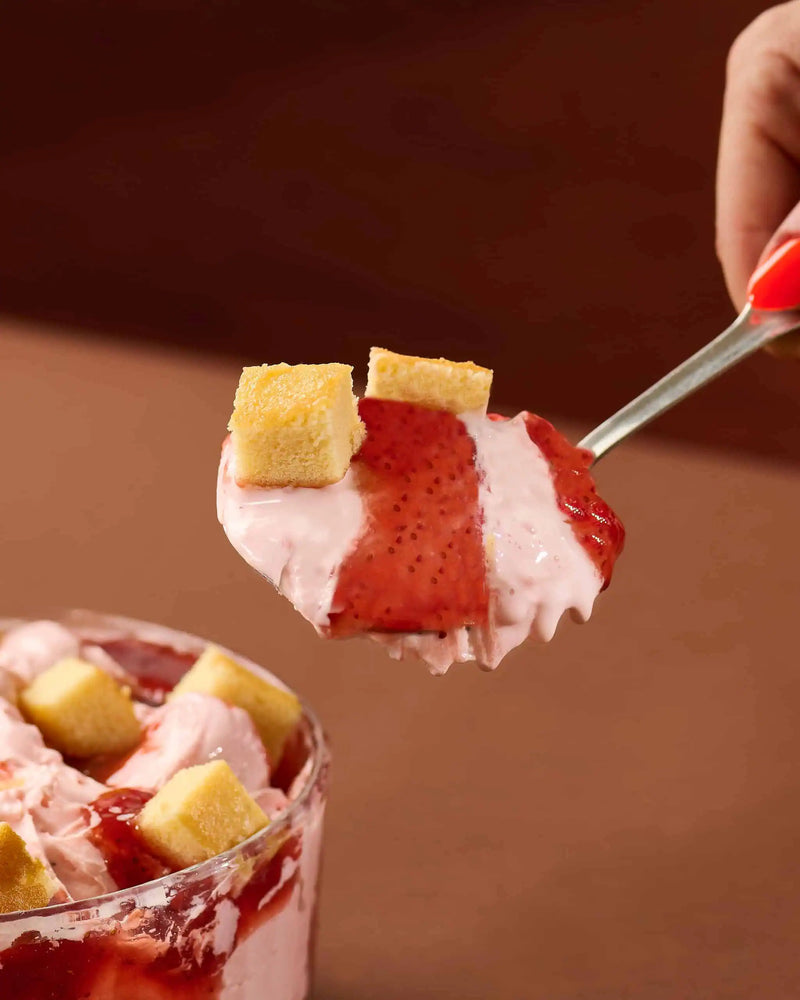 A close-up of a spoonful of strawberry ice cream dessert with red strawberry sauce, pink whipped cream, and small yellow cake cubes, held over a glass bowl containing more of the dessert.