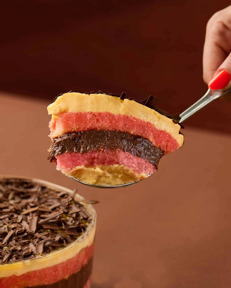 A close-up of a spoonful of layered dessert with pink sponge, dark brown chocolate, pale yellow cream, and caramel-like topping, with a hand holding the spoon and a matching dessert cup in the foreground.