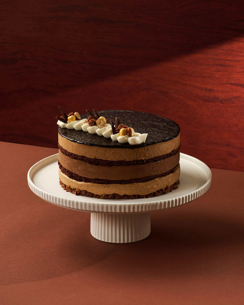 A chocolate-layer cake with white cream and nut toppings sits on a white pedestal cake stand against a warm brown studio background.