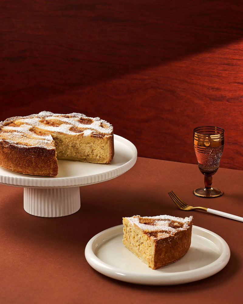 A slice of golden-brown cake dusted with powdered sugar sits on a small white plate in the foreground, with the remainder of the cake on a white pedestal stand behind it. A gold-toned goblet and fork are placed on a warm brown surface against a dark wood background.