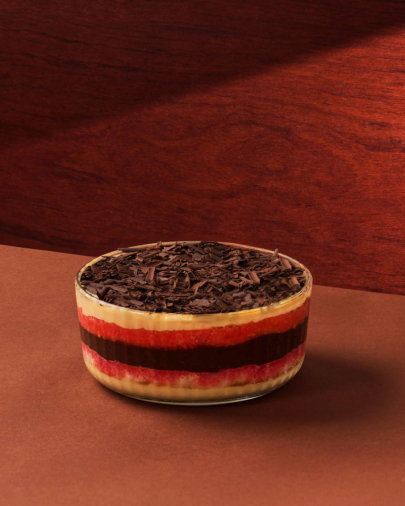 A layered dessert in a clear glass bowl, topped with dark chocolate shavings, set against a warm brown background.