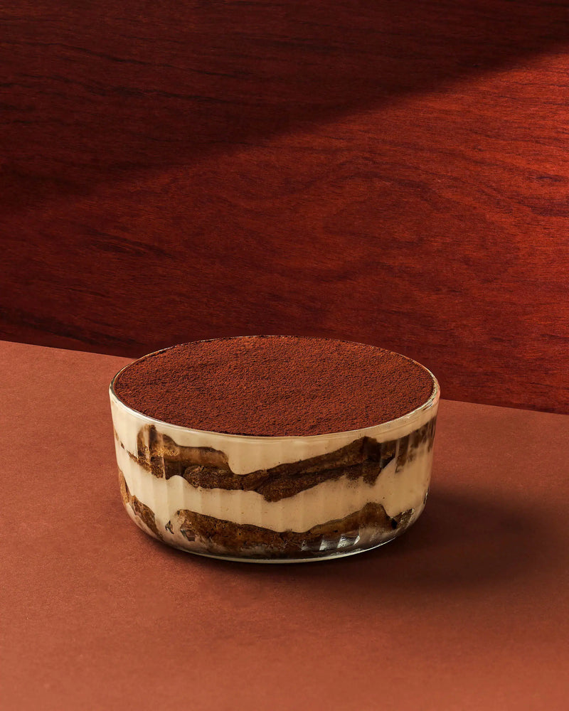 A layered dessert in a clear glass cup, topped with cocoa powder, set against a warm brown and terracotta background.