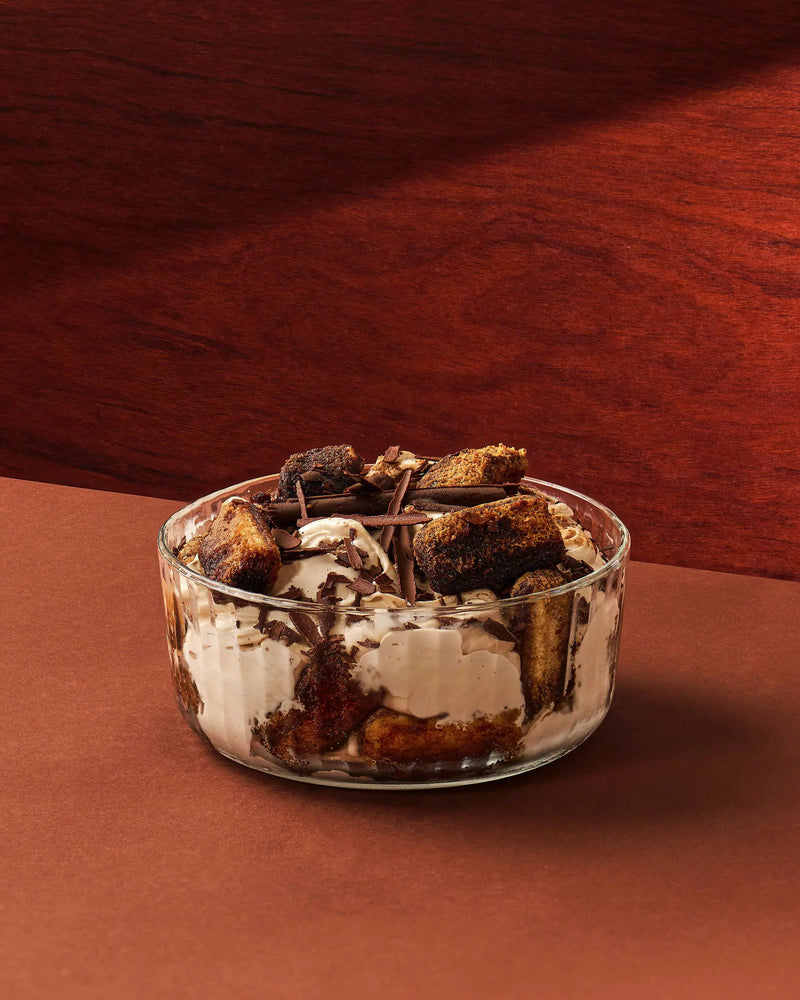 A clear glass bowl filled with a dessert layered in cream and dark chocolate, topped with chocolate chunks and chocolate shavings, set on a warm brown surface against a dark wood background.