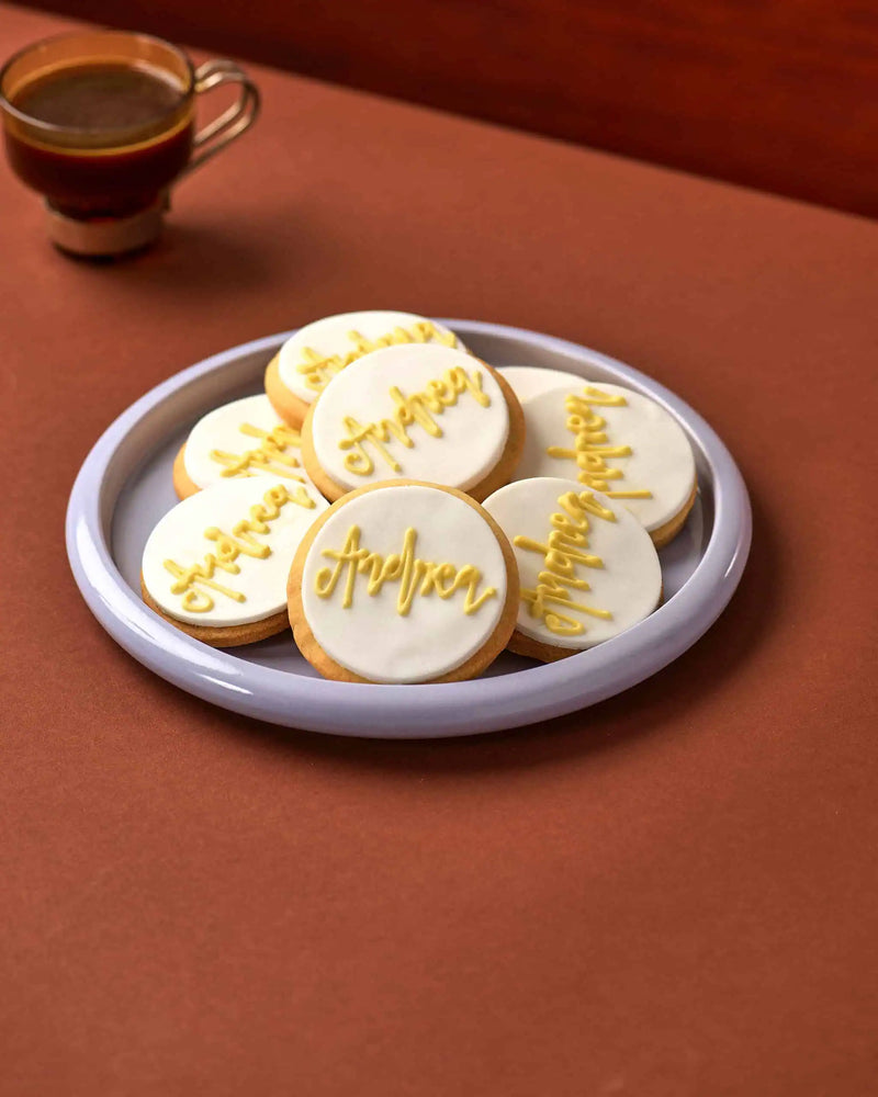A plate of round iced cookies with white icing and gold script names, set on a warm brown surface with a cup of coffee blurred in the background.