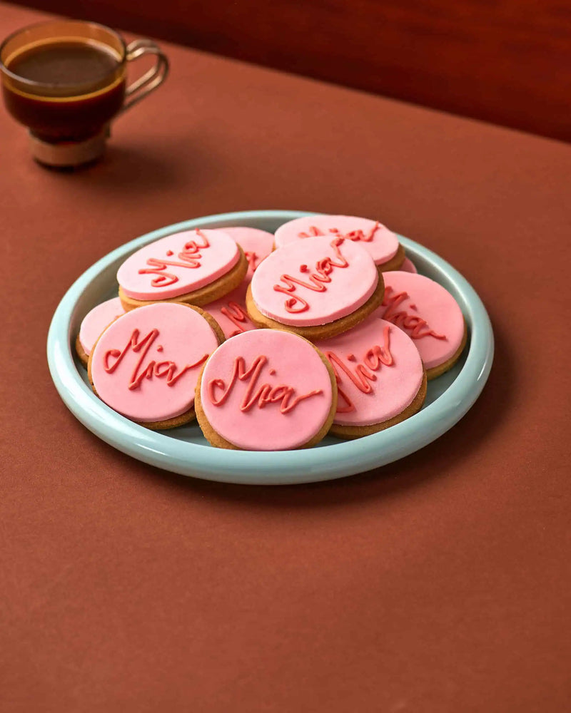 A plate of round pink frosted cookies with red script writing on top, set on a warm brown surface with a cup of coffee blurred in the background.